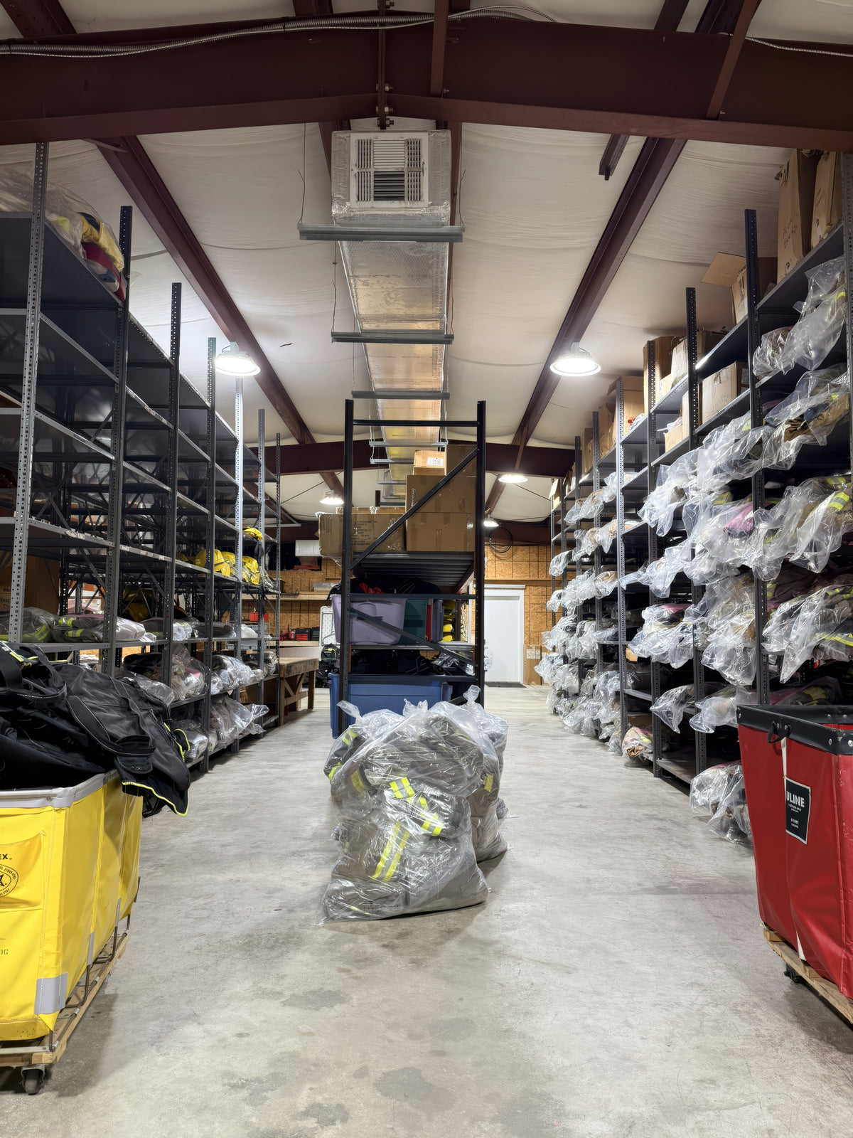 Looking down an aisle in Delta Fire & Safety's rental storage area, bagged turnout gear on steel shelving either side and canvas roller carts in the foreground.